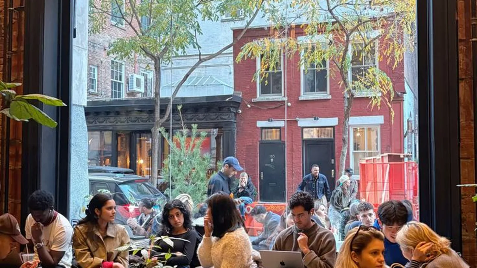 People are sitting inside a cozy, bustling cafe filled with large glass windows that overlook a city street.