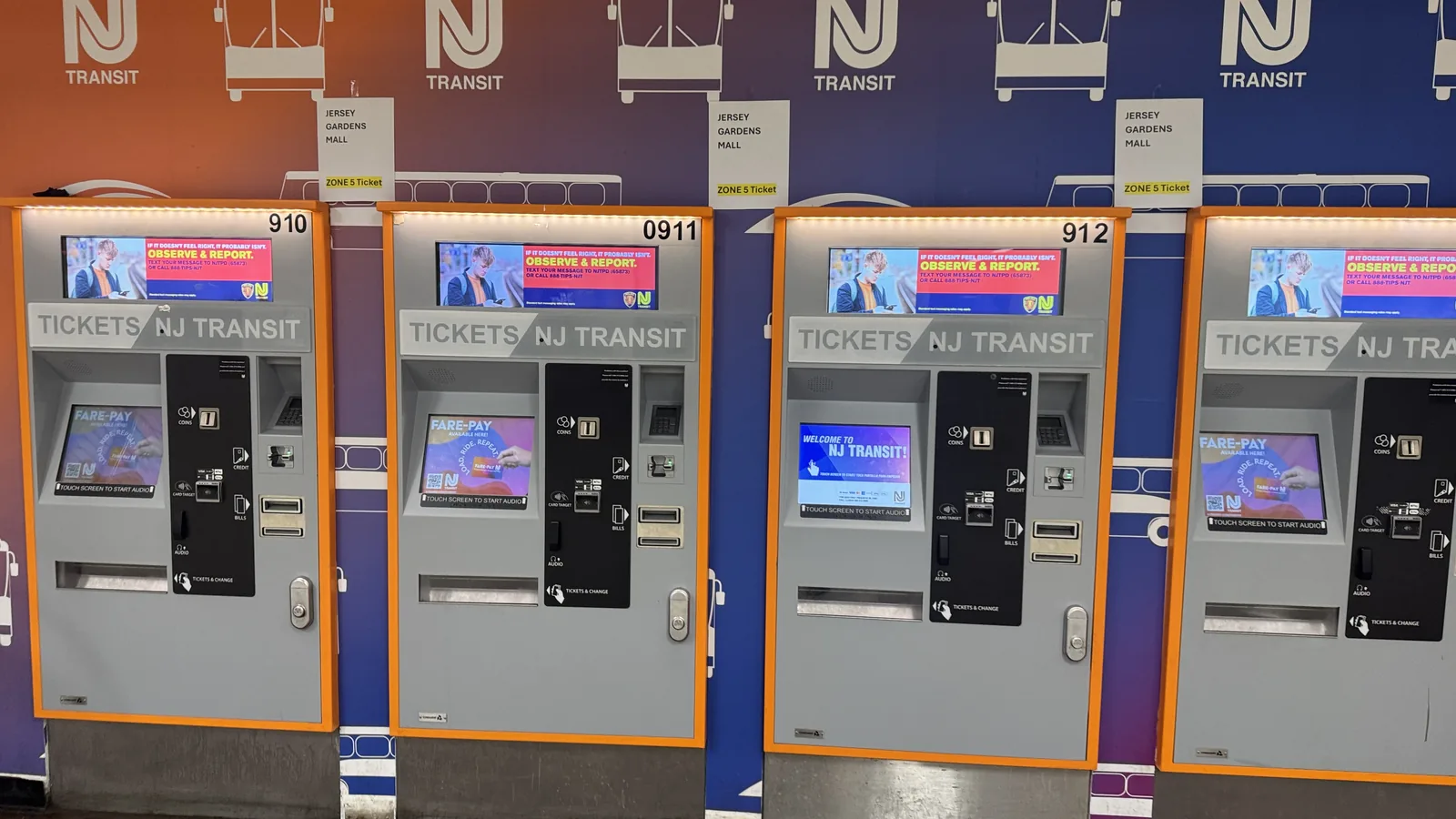 A row of four NJ Transit ticket vending machines stands against a brightly colored orange and blue wall.