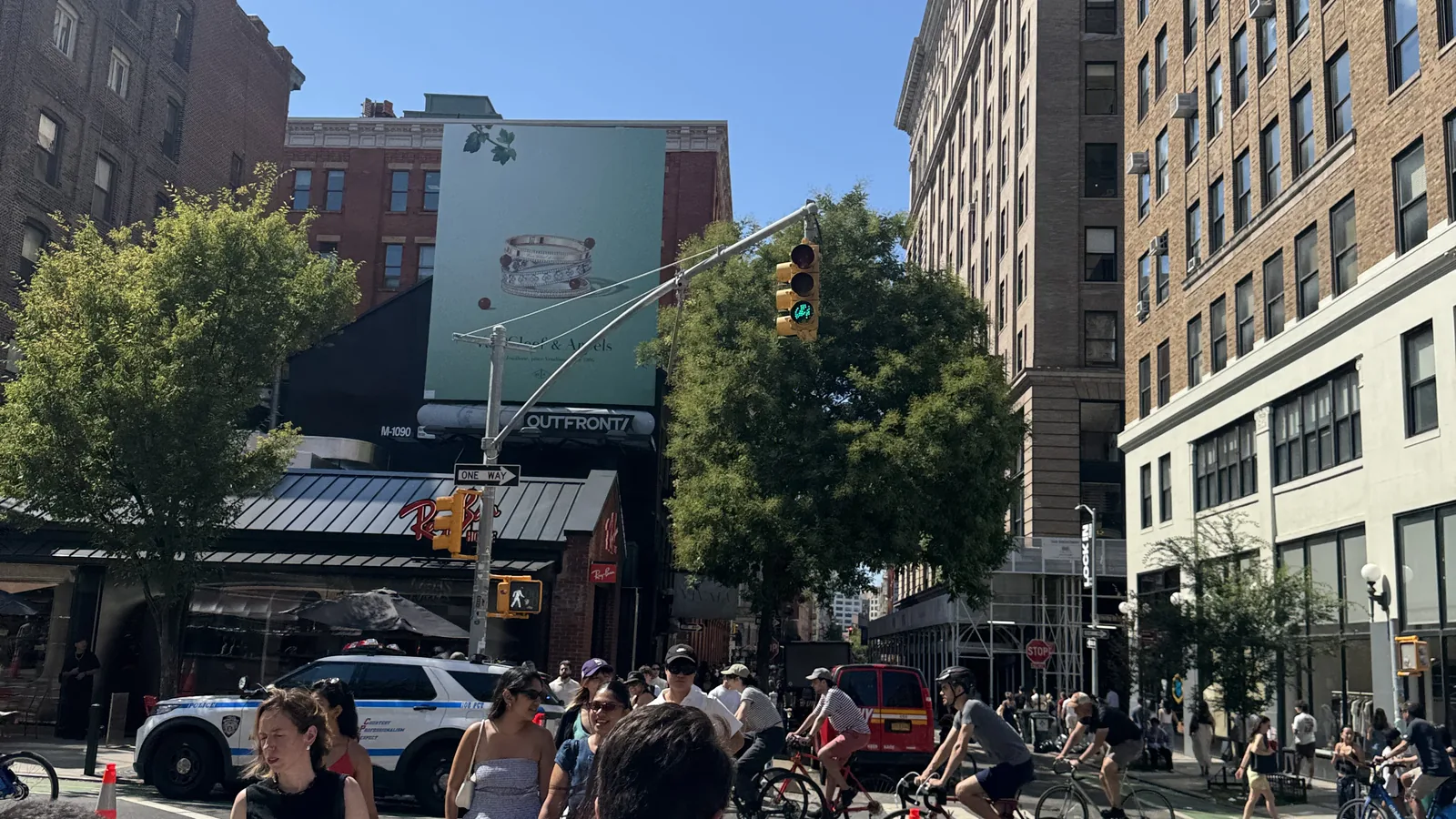 Pedestrians cross a busy city street while cyclists ride past under a bright, sunny sky.