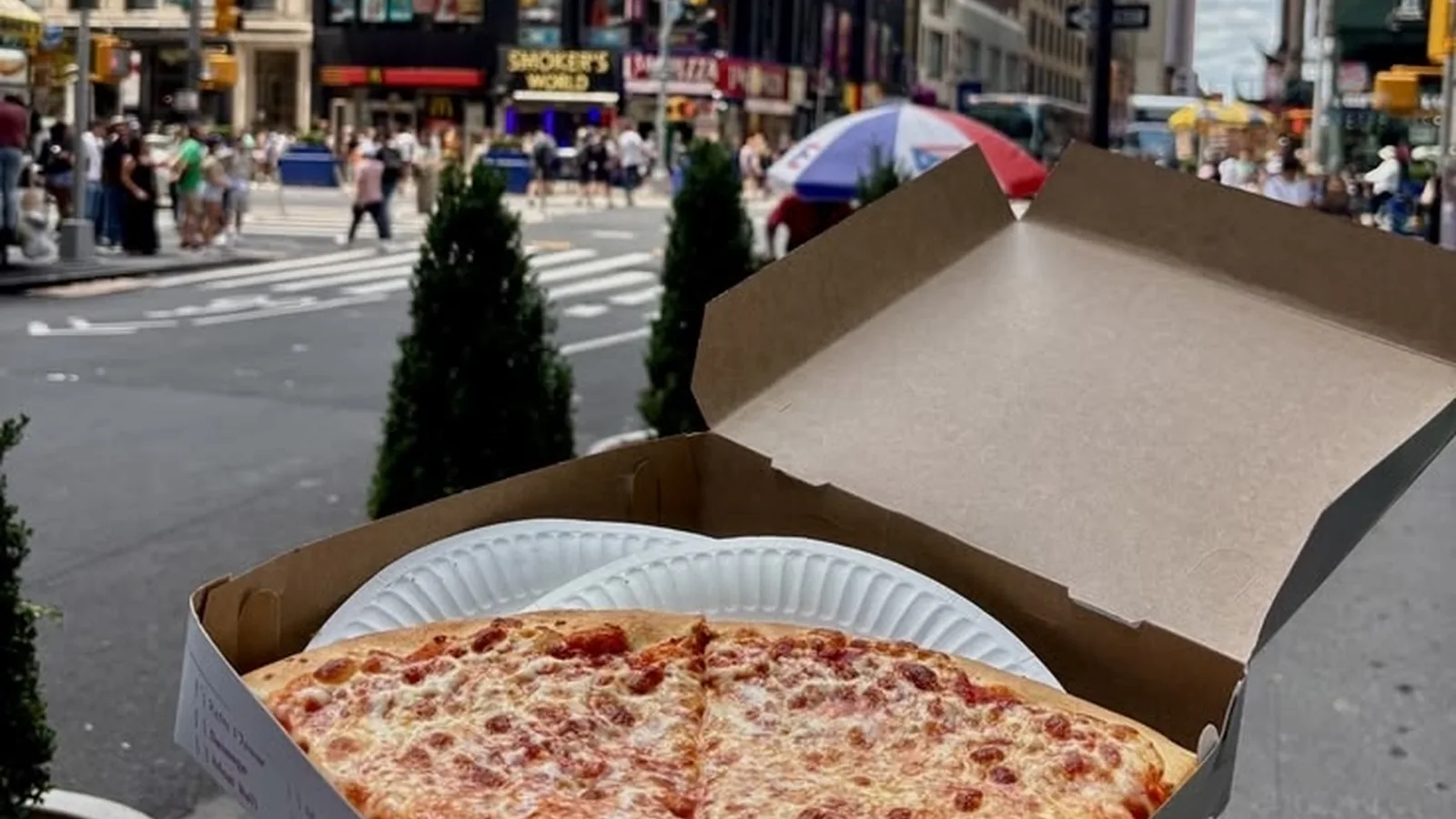 A New York street scene where two cheese pizza slices in a take-out box are held in the foreground, with the 'Smoker's World' store visible in the background