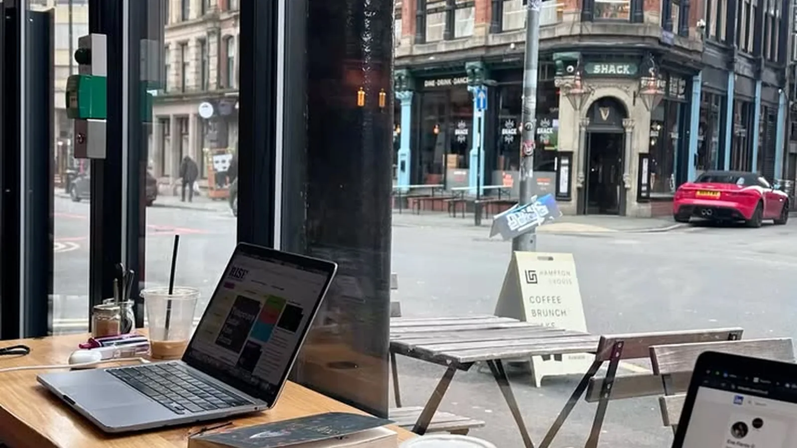 Laptops and coffee sit on a wooden counter inside a cafe looking out onto a European city street corner.