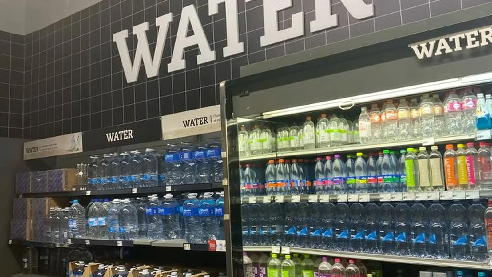 Bottled water is displayed on open shelves and in refrigerated coolers beneath a large "WATER" sign in a grocery store.
